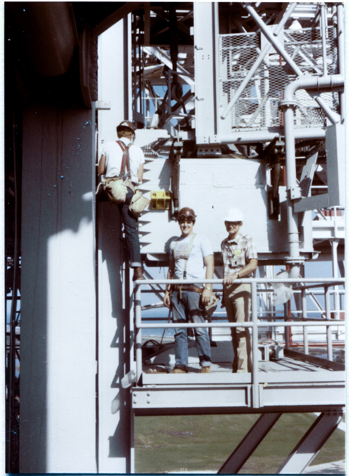 Image 135. OAA Lift 7 of 9. On the Orbiter Access Arm Lower Hinge Access Platform at Elevation 190'-6” on Side 1 of the Fixed Service Structure at Space Shuttle Launch Complex 39-B, Kennedy Space Center, Florida, we see Wade Ivey (right, facing the camera with the OAA's Lower Hinge Box behind him), owner of Ivey Steel Erectors, his son Kevin (center, facing the camera with the OAA's Lower Hinge Box behind him), and an unidentified Union Ironworker (facing away from the camera with his boot on the Platform handrail) working for Ivey Steel. The OAA is in the process of being bolted to its Support Strongback. Kevin was learning the craft of ironworking at the time, and went on to own and operate Ivey Construction (which remains in business to this day, and has expanded well beyond the realm of structural steel erection) after his father retired. Despite the fact that it was simply a means for Shuttle Flight Crews to walk out away from the FSS and enter the Vehicle prior to launch, the OAA was a exceedingly-complex thing, and you can get a glimpse of some of that complexity in this image. Photo by James MacLaren.